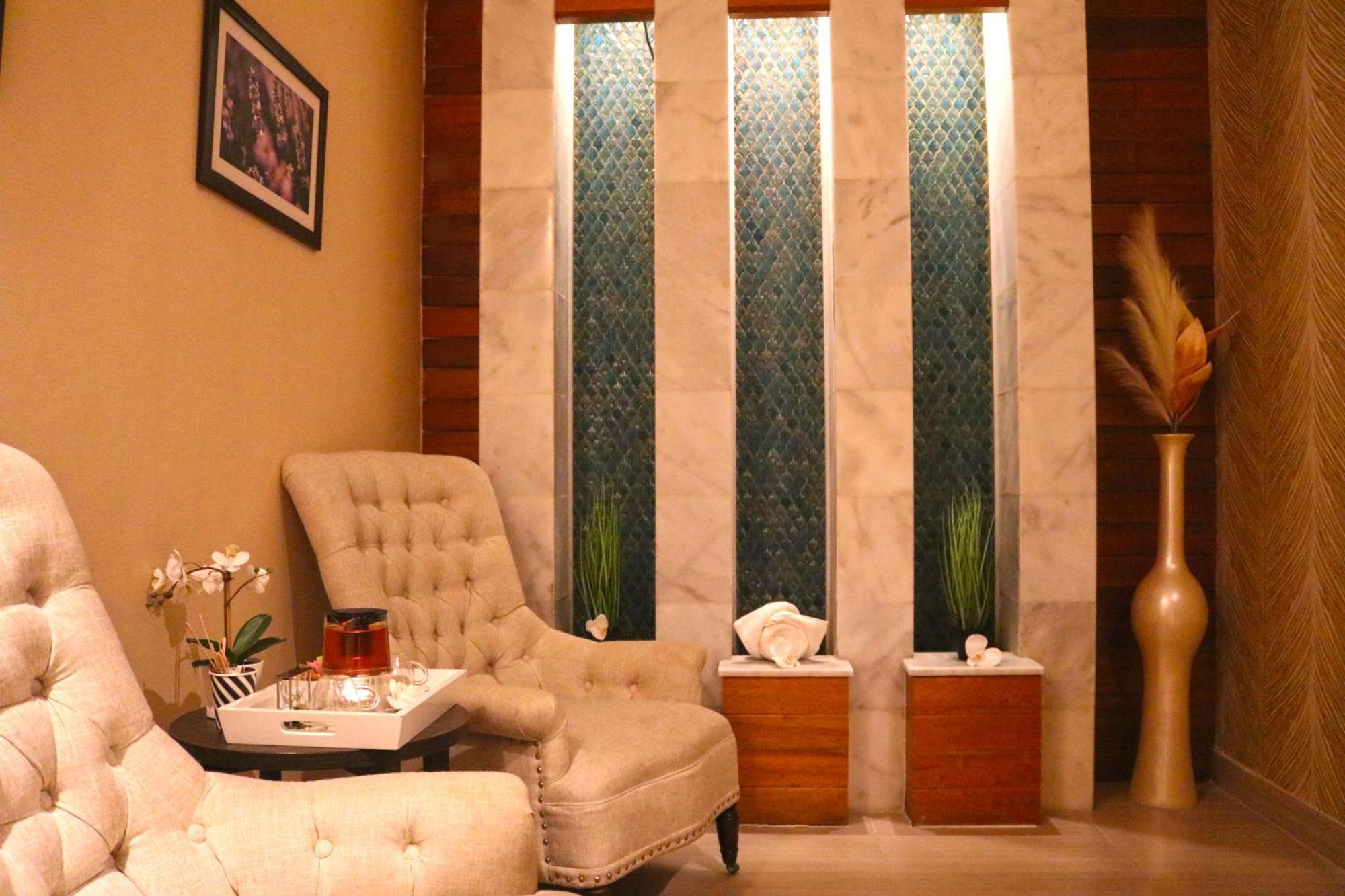 Lagoona Beach Luxury Resort And Spa Manama Dış mekan fotoğraf The image shows a relaxing interior space, likely a waiting area or lounge. There are two plush, beige armchairs positioned near a small table, which holds a glass pitcher and a cup. The background features tall, elegant windows with decorative blue 