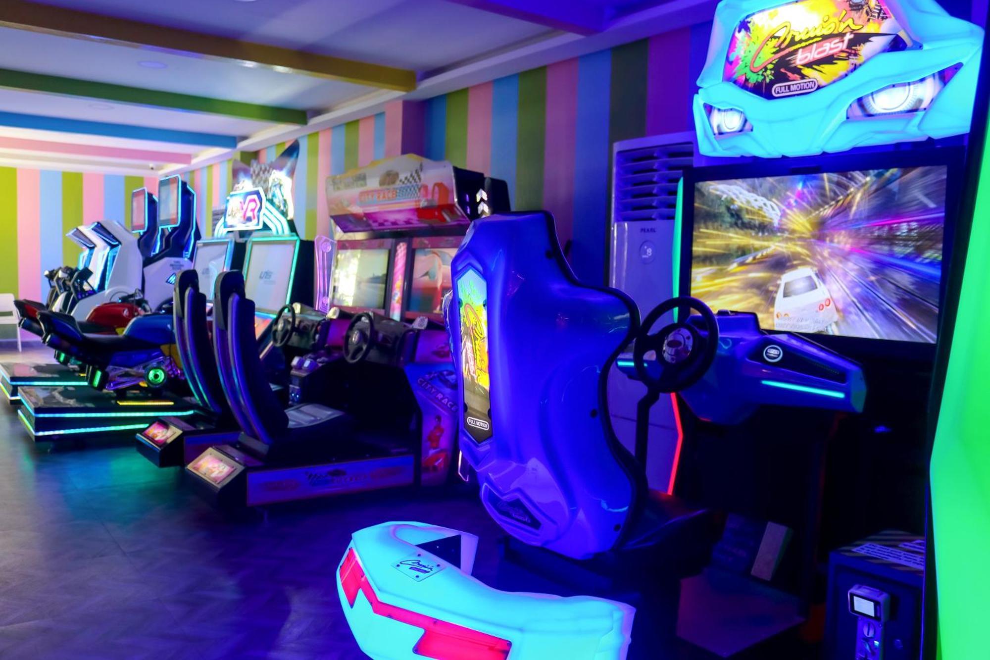 Lagoona Beach Luxury Resort And Spa Manama Dış mekan fotoğraf The photo shows an arcade area filled with various arcade machines. The setting is illuminated with colorful neon lights, creating a vibrant atmosphere. There are several racing game machines with steering wheels and seats, each accompanied by screen