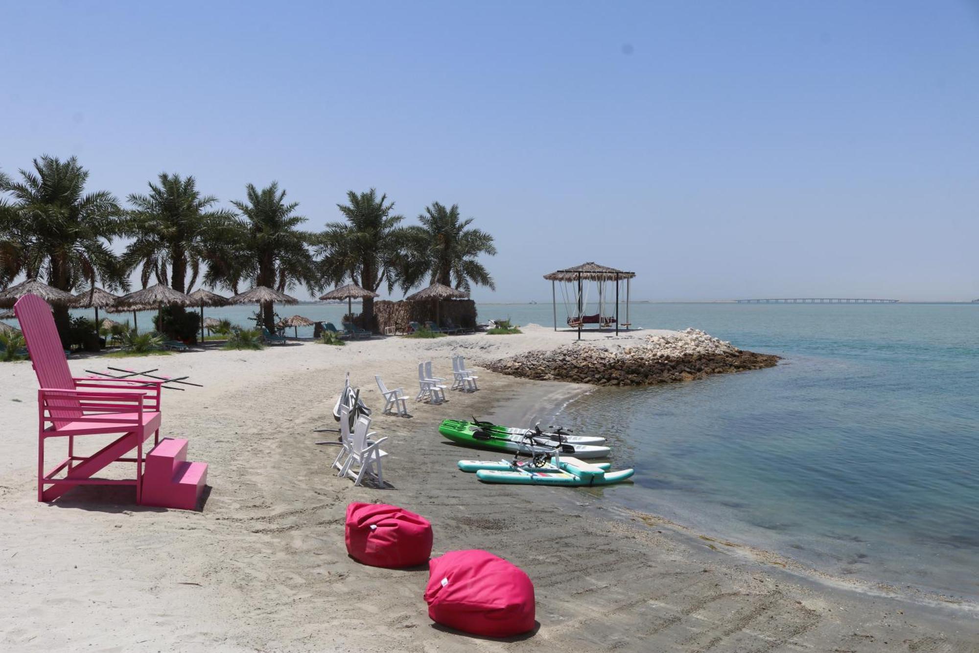 Lagoona Beach Luxury Resort And Spa Manama Dış mekan fotoğraf The photo depicts a serene beach scene with clear blue water and a sunny sky. In the foreground, there are colorful pink bean bags and a large pink chair on the sand. A few white chairs and loungers are positioned near the water's edge. To the side, 