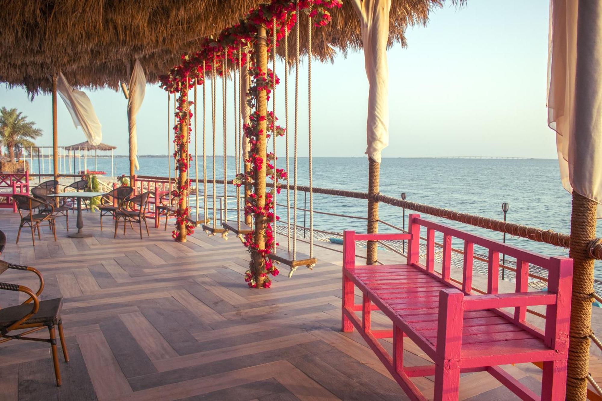 Lagoona Beach Luxury Resort And Spa Manama Dış mekan fotoğraf The photo depicts a scenic outdoor area by the water, with a wooden deck and a thatched roof. Two swings adorned with flowers are hanging from the ceiling, providing a charming touch to the space. In the foreground, there is a bright pink bench, addi