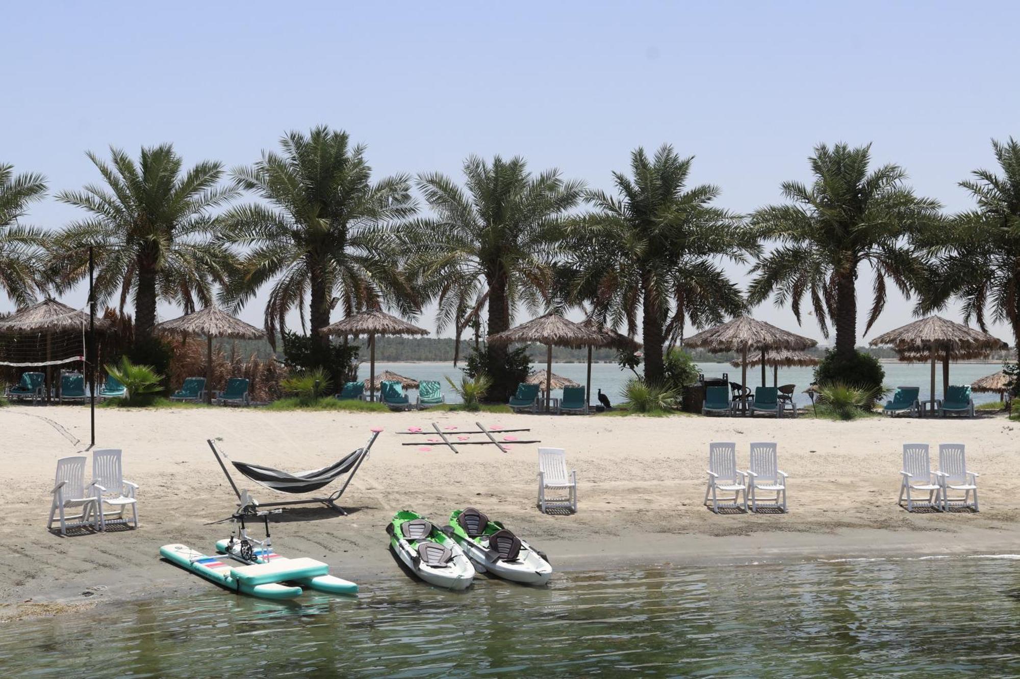 Lagoona Beach Luxury Resort And Spa Manama Dış mekan fotoğraf The photo depicts a serene beach scene with several palm trees lining the shore. There are thatched umbrellas providing shade, and a few lounge chairs are positioned on the sand. A hammock is set up between two supports, inviting relaxation. In the f