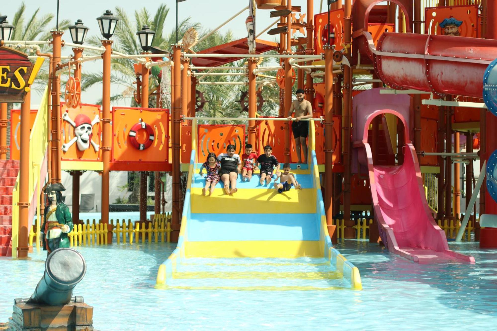 Lagoona Beach Luxury Resort And Spa Manama Dış mekan fotoğraf The photo shows a colorful water park scene featuring a play area with multiple slides. There are children enjoying themselves, sliding down bright yellow and pink slides into a pool of water. The structure has pirate-themed decorations, including a 