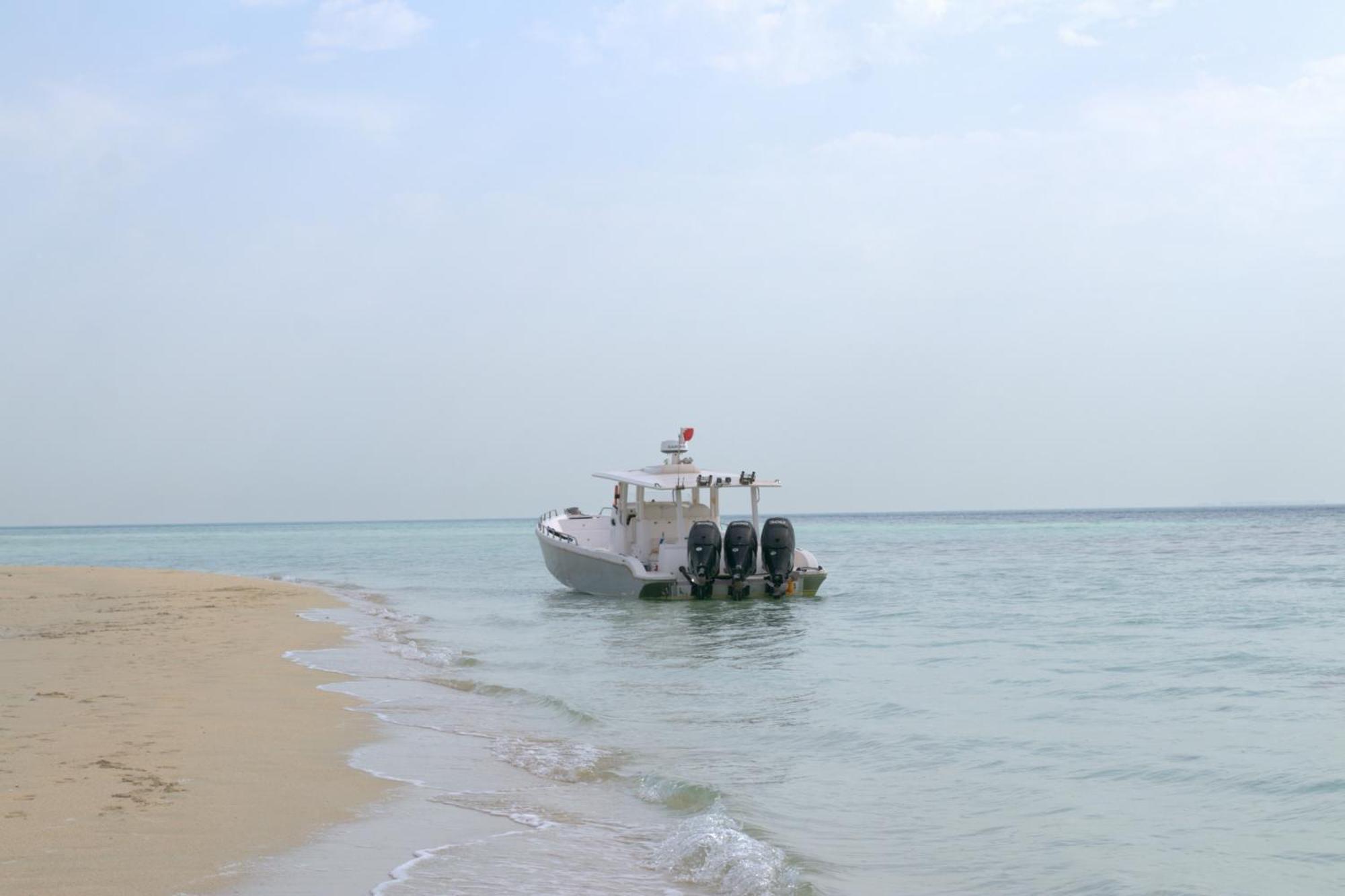 Lagoona Beach Luxury Resort And Spa Manama Dış mekan fotoğraf The photo depicts a small boat anchored near a sandy beach, partially submerged in calm, clear water. The boat is white and features three outboard motors on its stern. In the background, the sea stretches out toward the horizon under a light, cloudy