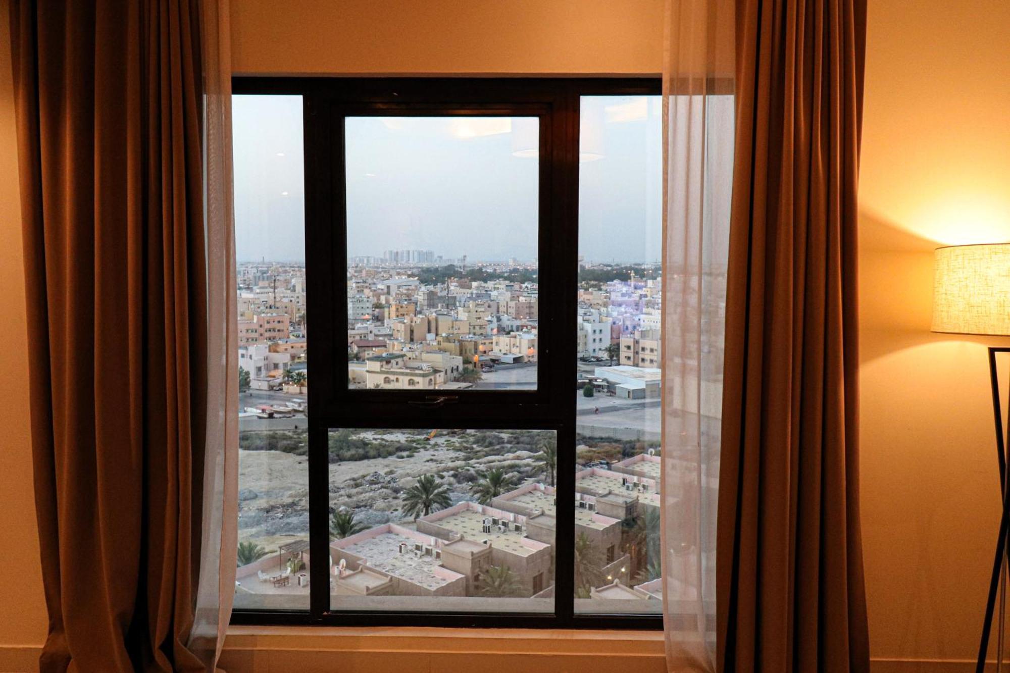Lagoona Beach Luxury Resort And Spa Manama Dış mekan fotoğraf The photo shows a window with a black frame and sheer curtains. Through the window, there is a view of a cityscape, displaying a densely populated area with various buildings and rooftops. The sky appears slightly overcast, suggesting a muted lightin