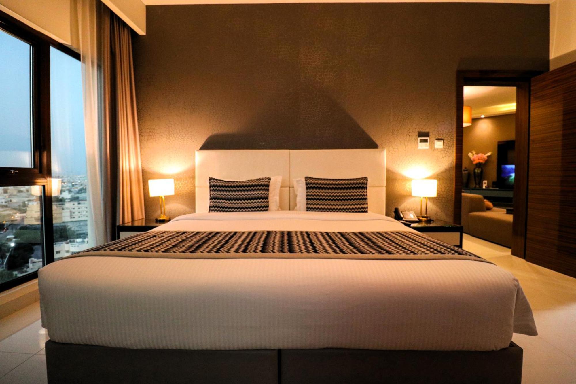 Lagoona Beach Luxury Resort And Spa Manama Dış mekan fotoğraf The photo depicts a modern hotel room featuring a large bed with a white comforter and two decorative pillows. The headboard is light-colored, and the walls are painted a dark hue. On either side of the bed, there are bedside tables with lamps. The r