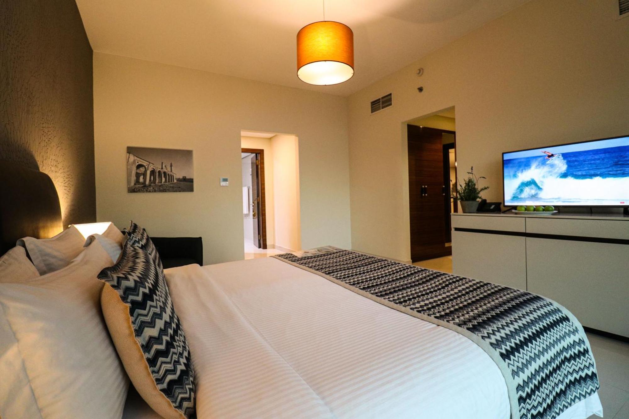 Lagoona Beach Luxury Resort And Spa Manama Dış mekan fotoğraf The photo shows a well-decorated hotel room. There is a large bed with white linens and decorative pillows featuring a zigzag pattern. On the wall beside the bed is a piece of artwork, and there is a modern television on a white furniture unit opposi