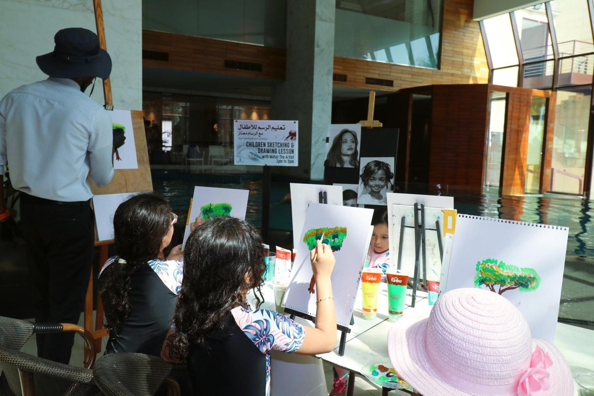 Lagoona Beach Luxury Resort And Spa Manama Dış mekan fotoğraf The photo shows a group of children and an adult engaged in an art activity near a pool area. The children are seated at a table, painting on canvases or paper, with a focus on trees, as indicated by the green paint they are using. An adult, wearing 