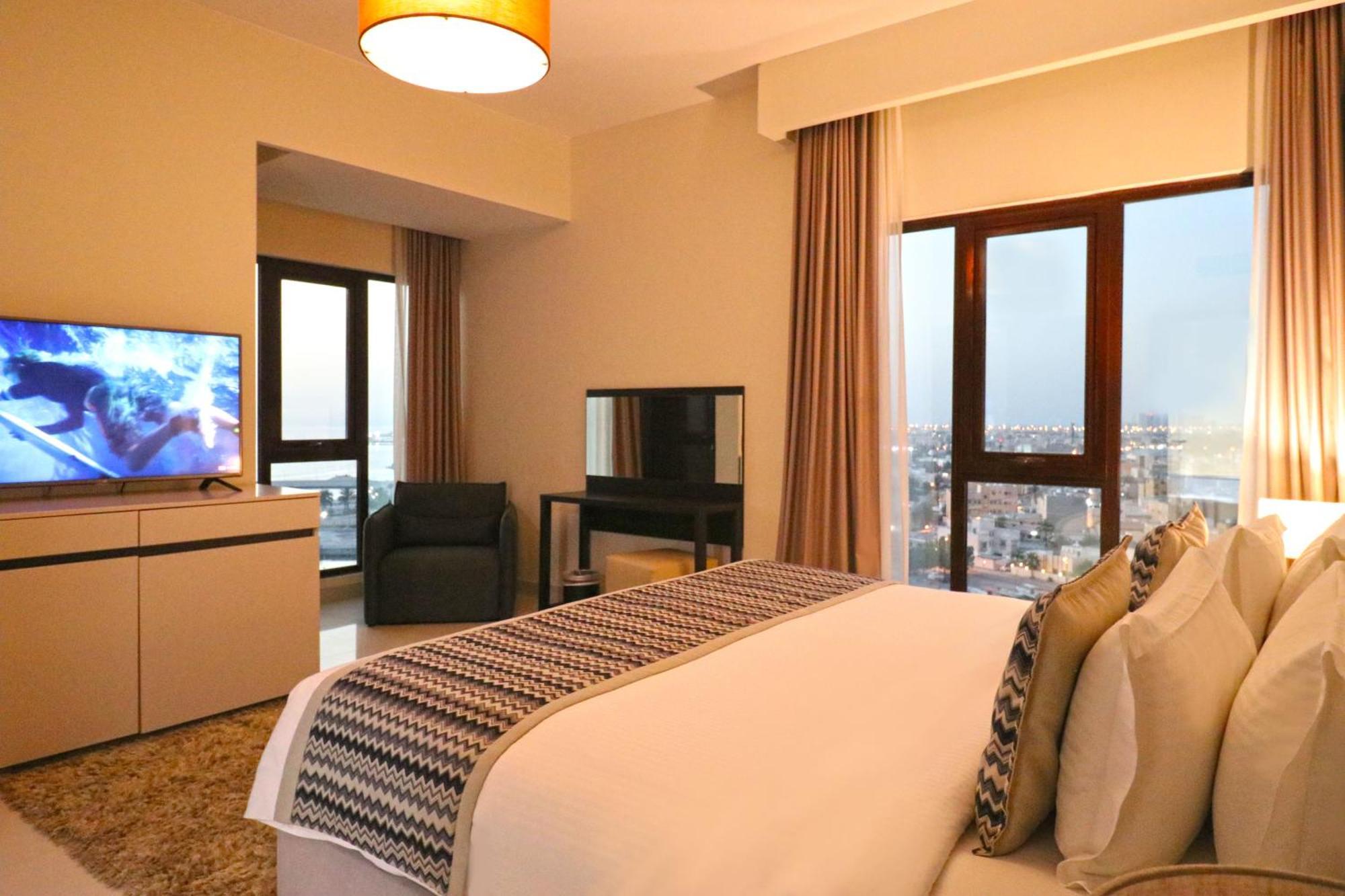 Lagoona Beach Luxury Resort And Spa Manama Dış mekan fotoğraf The photo shows a modern hotel room or suite. It features a neatly made bed with decorative pillows in the foreground. To the left, there is a TV on a cabinet, and further back, a comfortable chair is situated in front of a large window that likely o