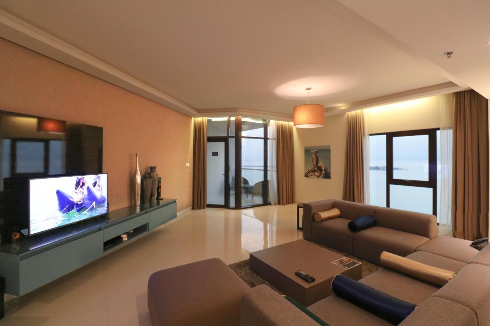 Lagoona Beach Luxury Resort And Spa Manama Dış mekan fotoğraf The photo shows a modern living room with a sleek design. There is a large wall-mounted television on a contemporary entertainment unit, with decorative items placed beside it. The room features a comfortable sectional sofa in light tones, with a cof