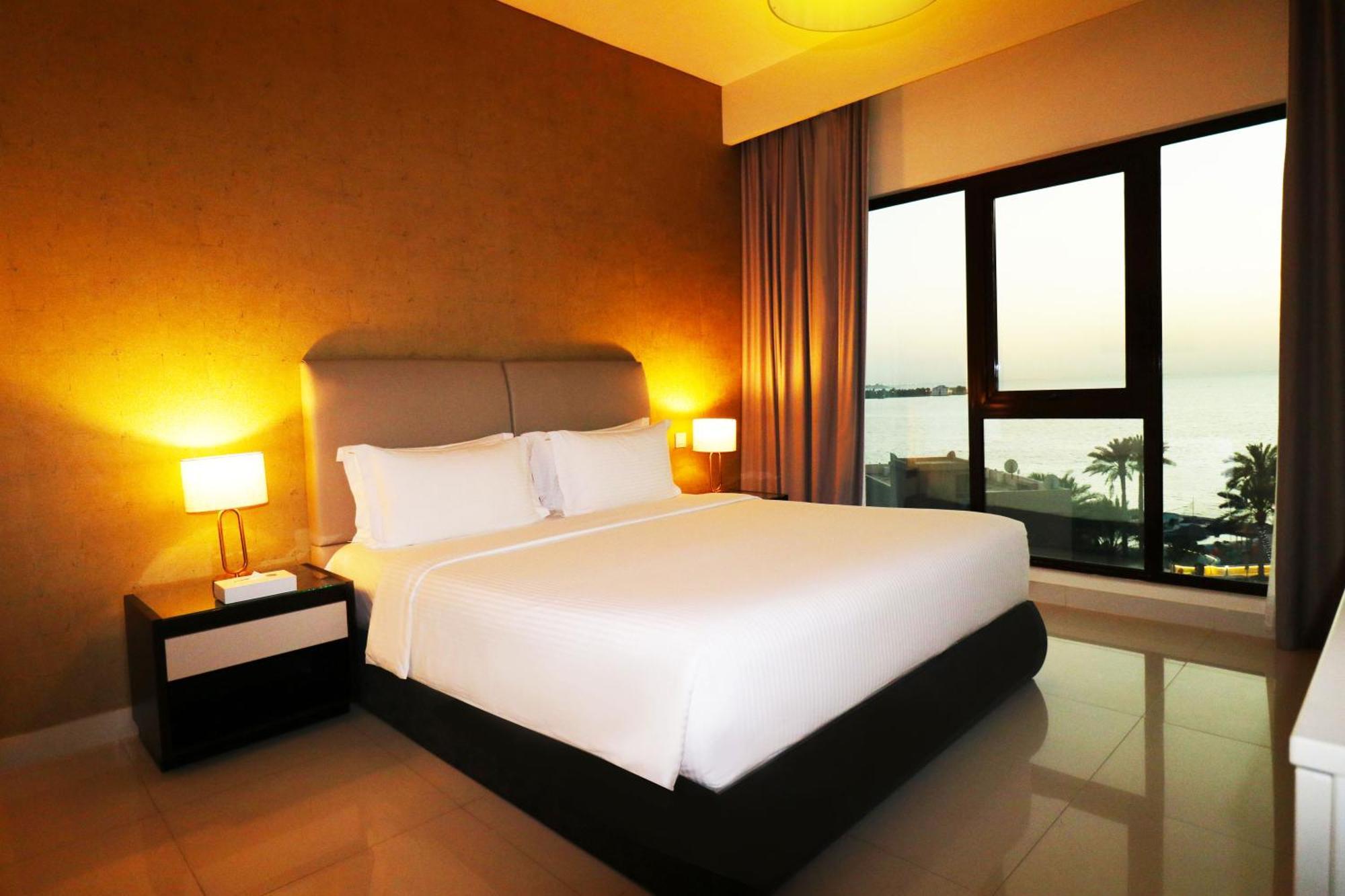 Lagoona Beach Luxury Resort And Spa Manama Dış mekan fotoğraf The photo shows a modern hotel bedroom with a large, comfortable bed dressed in white linens. The room features warm lighting, including bedside lamps, creating a cozy atmosphere. There are large windows with a view of the outdoors, possibly overlook