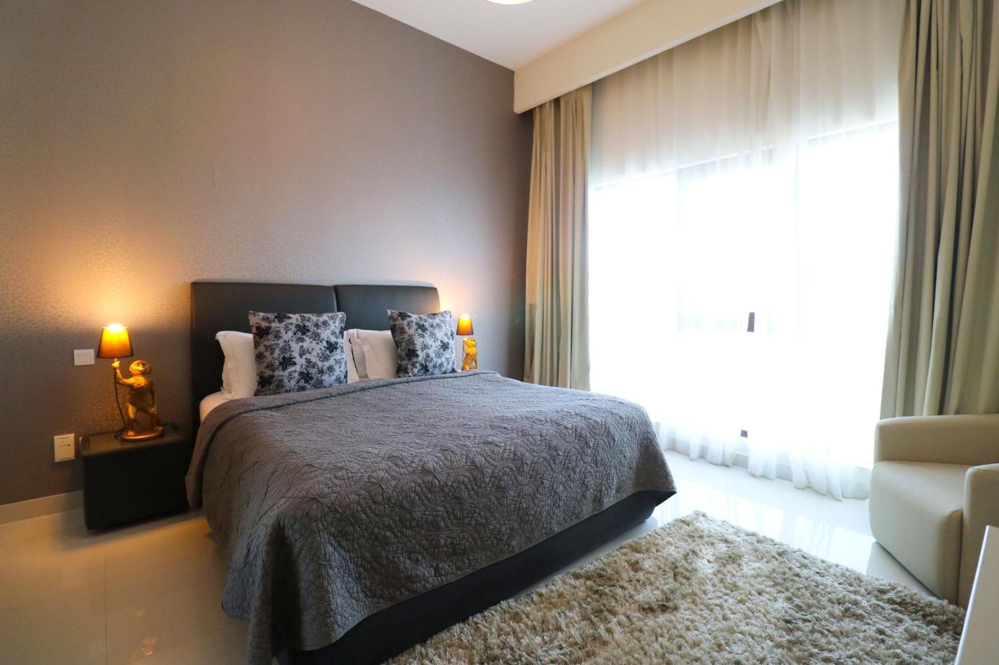 Lagoona Beach Luxury Resort And Spa Manama Dış mekan fotoğraf The photo shows a stylish bedroom with a modern design. It features a large bed with a dark gray bedspread and decorative pillows. On either side of the bed are two wooden nightstands, each with a lamp. There is a large window covered with light, flo